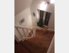 Holztreppe in einem Wohnhaus mit weiß gestrichenen Wänden und einem Fenster mit weißen Rahmen. Neben dem Fenster steht eine grüne Zimmerpflanze, an den Wänden hängen zwei kleine Bilder, und eine Wandlampe beleuchtet den Bereich.