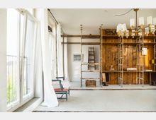 Heller, minimalistischer Innenraum mit großen, geöffneten Fenstern und weißen Vorhängen. Holzregale mit Metallrahmen bedecken eine Wand, daneben befinden sich eine Leiter, ein Sessel und ein Kronleuchter mit weißen Lampenschirmen.