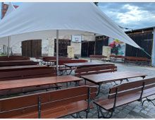 Offener Hofbereich mit Kopfsteinpflaster und langen Holzbänken und -tischen, teilweise überdacht von einem weißen Sonnensegel. Im Hintergrund sind Holztore, bemalte Wände und eine alte Basketballkorbanlage zu sehen.