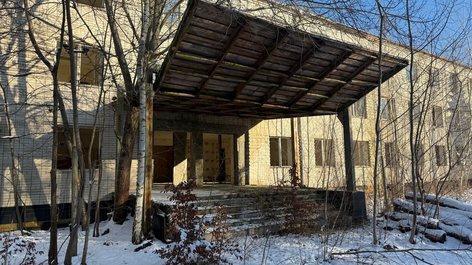 Eingang eines verlassenen, zweigeschossigen Backsteingebäudes mit breiter Treppe und einem großen, aus Holzplanken gefertigten Vordach auf Stahlstützen. Davor stehen kahle Bäume und Sträucher in einer verschneiten Außenanlage, mehrere Fenster sind ohne Verglasung.