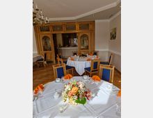 Ein festlich gedeckter Raum mit runden Tischen, weißen Tischdecken, blauen Polsterstühlen und orangefarbenen Servietten. Im Vordergrund steht ein Tisch mit einem Blumenarrangement, im Hintergrund sind Holztüren mit Glaseinsätzen und eine kleine Theke sichtbar.