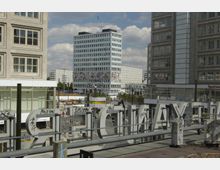 Städtischer Platz mit einem hohen Bürogebäude im Hintergrund, das eine auffällige Wandkunst mit bunten Motiven zeigt. Im Vordergrund sind große, mit Graffiti versehene Buchstaben und umliegende moderne Gebäude zu sehen.