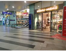 Innenansicht eines Bahnhofsgebäudes mit Rolltreppen, Geschäftseingängen und Beschilderung. Im Vordergrund befinden sich Geschäfte wie ein Nanu-Nana, ein McDonald's und Rossmann, sowie Wegweiser zu den Gleisen 3-4 und 11-12.