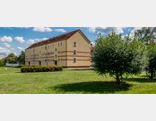 Mehrstöckiges Wohngebäude mit gelbem Putz, dunklem Satteldach und mehreren rechteckigen Fenstern, umgeben von einer Grünfläche mit Bäumen, Büschen und einem Wäschetrockenplatz. Der Himmel ist teils bewölkt mit viel Sonnenschein.