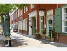 Straßenansicht mit historischen Ziegelgebäuden und weiß gerahmten Fenstern, vor denen Tische mit grünen Sonnenschirmen und Blumenkörben auf einem gepflasterten Gehweg stehen; Laden- und Café-Schilder sind an den Fassaden angebracht. Ein schattenspendender Baum säumt den Gehweg.