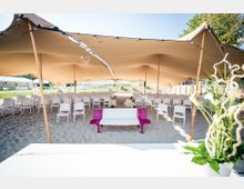 Zeltüberdachter Veranstaltungsbereich auf einem Sandboden mit weißen Stühlen, die in Reihen angeordnet sind, und einer weiß-pinken Bank in der Mitte; im Vordergrund Pflanzen und im Hintergrund offene Grünflächen.