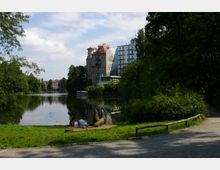 Ein Park mit einem See, umgeben von Bäumen und vereinzelten Gebäuden mit heller Fassade im Hintergrund. Im Vordergrund befindet sich eine Wiese mit einer Skulptur und zwei Personen, die am Ufer sitzen.