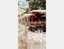 Tisch in einem Außenbereich mit Weingläsern und einem Glasgefäß voller weißer Herz-Dekorationen im Vordergrund, umgeben von Sonnenschirmen, Holzmöbeln und Bäumen im Hintergrund.