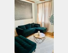 Wohnzimmer mit dunkelgrünem, gepolstertem Ecksofa und passendem Hocker auf hellem Teppich, davor ein achteckiger Couchtisch mit Holzplatte und Metallgestell. An der Wand hängt ein großes Periodensystem-Poster; daneben ein Fenster mit bodenlangen, beigen Vorhängen und Holzboden sowie eine Zimmerpflanze in der Ecke.