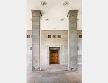 Überdachter Eingangsbereich eines Steingebäudes mit zwei massiven, rechteckigen Säulen und einer hellen Decke mit eingelassenen Rundlampen. Mittig befindet sich eine hölzerne Doppeltür, darüber und seitlich kleine vergitterte Fensteröffnungen in der Steinwand.