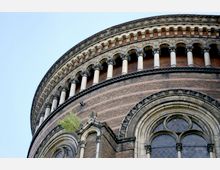 Runde Backsteinfassade eines historischen Gebäudes mit dekorativen Bögen, Säulen und großen gotischen Fenstern mit bleigefasstem Glas, umgeben von detaillierten steinernen Ornamenten. Ein kleiner Baum wächst aus der Fassade.