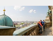 Menschen stehen auf einer Aussichtsplattform mit Geländer und blicken über die Stadt Berlin. Im Vordergrund ist eine grüne Kuppel zu sehen, im Hintergrund sind historische Gebäude, darunter der Berliner Dom, sowie eine weite Stadtlandschaft mit vereinzelten Wolken am Himmel.