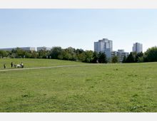Weitläufige Grünfläche in einem Stadtpark mit einzelnen Spaziergängern und Personen auf einer Bank; im Hintergrund moderne Wohngebäude und dicht bewachsener Baumbestand.