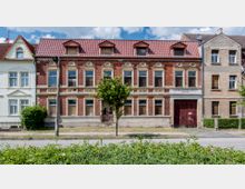 Fassade eines alten, zweistöckigen Backsteingebäudes mit roten Dachziegeln und mehreren symmetrisch angeordneten Fenstern, teilweise mit verschlossenen Fensterläden. Das Gebäude ist von Nachbarhäusern flankiert und liegt an einer Straße mit Gehweg und Grünstreifen im Vordergrund.