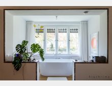 Helles Badezimmer mit einer freistehenden, weißen Badewanne vor einem großen Fenster mit Vorhängen. Links eine grüne Monstera-Pflanze, rechts ein gerahmtes Bild mit roten Akzenten.