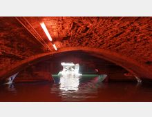 Backsteinbogen einer niedrigen Brücke über einem Wasserkanal, beleuchtet durch ein rotes Licht. Im Hintergrund ist ein Ausgang mit Tageslicht und reflektierendem Wasser sichtbar.