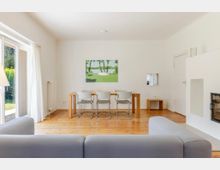 Helles Wohn- und Esszimmer mit Holzboden, großer Terrassentür mit Vorhängen links und einem Esstisch mit vier Stühlen unter einer Pendelleuchte. Rechts befindet sich ein weißer Kamin mit Nische, an der Rückwand hängt ein Landschaftsbild.