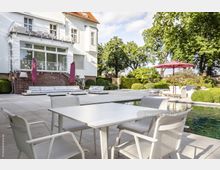 Großer moderner Garten mit Terrasse, die mit hellen Steinplatten belegt ist, Sitzmöbeln und Sonnenschirmen. Im Hintergrund ein weißes Gebäude mit roten Dachziegeln und schmiedeeisernen Balkongeländern, umgeben von Bäumen und gepflegten Hecken.