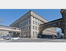 Eckansicht eines großen, historischen Verwaltungsgebäudes mit einer Fassade aus hellem Stein und zahlreichen Fenstern. Ein steinerner Bogen verbindet zwei Gebäudeteile, darunter verläuft eine Straße; daneben ist eine moderne Fußgängerbrücke aus Glas und Metall zu sehen.