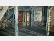 Ein rustikaler Dachbodenraum mit sichtbaren Holzbalken und Steinwänden. Im Vordergrund stehen Pinsel in einem Ständer, an der Wand hängt ein Gemälde eines Gesichts, und eine offene Tür führt zu einem Raum mit abstrakten Kunstwerken im Hintergrund.