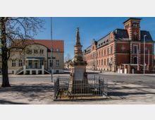 Städtischer Platz mit einem historischen Sandsteinbrunnen im Vordergrund, umgeben von einem schmiedeeisernen Zaun. Im Hintergrund befinden sich ein Gebäude mit rotem Ziegeldach und ein größeres Ziegelbauwerk mit Ornamenten und Dachgauben.
