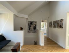 Ein heller, moderner Flur mit Dachschräge, weiß gestrichenen Wänden und hellem Fischgrät-Holzboden. An den Wänden hängen mehrere abstrakte Gemälde, und am Ende des Flurs befindet sich eine Tür, die zu einem Raum mit großen Fenstern führt.