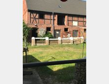 Altes Backstein-Fachwerkgebäude mit geneigtem Ziegeldach, mehreren Holz­türen und kleinen Fenstern, teils mit beschädigtem Putz. Davor liegt eine Rasenfläche mit einem niedrigen Zaun aus Steinpfeilern und Holzlatten.