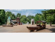 Ein Spielplatz mit Sandboden, umgeben von Bäumen, zeigt mehrere fantasievoll gestaltete Spielgeräte, darunter eine Holzbootstruktur, eine Rutsche in Form eines Meerjungfrauenkörpers und bunte Spielhäuschen. Im Hintergrund sind spielende Kinder und Erwachsene sowie Grünflächen und ein kleiner Teich sichtbar.