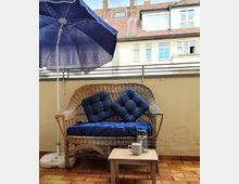 Kleine Terrasse mit einem geflochtenen Sofa mit blauen Kissen, einem kleinen Beistelltisch mit einer Tasse und einer French Press sowie einem blauen Sonnenschirm. Im Hintergrund sind Häuser mit Ziegeldächern und Fenstern zu sehen.