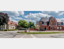 Straßenszene mit einem historischen Backsteingebäude auf der rechten Seite, das ein Restaurant beherbergt, und Wohnhäusern mit roten Ziegeldächern auf der linken Seite. Vor dem Gebäude befinden sich Schilder mit Werbungen, umgeben von Bäumen und Straßenkreuzungen.