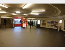 Eine unterirdische Passagenhalle in einem U-Bahn-Bereich mit beleuchteter Decke. Links eine Treppe, in der Mitte ein rot beleuchteter Bäckereistand, rechts Wanddekoration mit historischen Fotografien und einem Bildschirm.