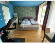 Schlafzimmer mit Doppelbett vor einer Wand mit tropischer Tapete. Der Raum hat blaue Wände, einen Holzboden, bodentiefe Fenster mit weißen Vorhängen und einen Heimtrainer im Vordergrund.