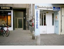 Straßenansicht mit einem Café links, erkennbar durch das leuchtende "Coffee"-Schild, einem Eingang mit Glastür in der Mitte und einem Geschäft für gebrauchte Haushaltsgeräte rechts mit der Aufschrift „Haushaltsgeräte An & Verkauf“. Zwei Fahrräder stehen vor dem Café, die Umgebung ist mit Kopfsteinpflaster versehen.