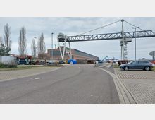 Industriegebiet mit einer großen Backsteinhalle im Hintergrund, daneben ein Kran- und Lagerbereich mit Stapeln von Baumaterialien. Im Vordergrund führt eine asphaltierte Straße entlang eines Parkplatzes mit einem geparkten Auto und einzelnen Bäumen am Rand.