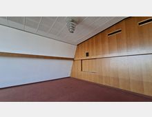 Leerer Innenraum mit rotem Teppichboden, weißer Wandfläche und großer Holzvertäfelung an der rechten Seite mit eingelassenen Lüftungsöffnungen und einer schmalen Ablage. An der Decke befinden sich perforierte Akustikplatten und eine zentrale Deckenleuchte.