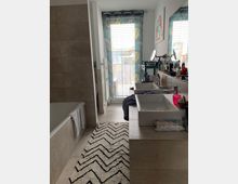 Modernes Badezimmer mit beigen Fliesen, einer Badewanne links, zwei weißen Waschbecken auf einer Steinablage rechts und einem großen Fenster mit einem Vorhang mit Pflanzenmuster im Hintergrund. Am Boden liegt ein Teppich mit Zickzack-Muster.