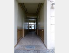 Schmaler, überdachter Durchgang mit cremefarbenen und braunen Wänden, die mit einer Leistenverzierung versehen sind, sowie einem gepflasterten Boden, der zu einer gläsernen Tür mit Sprossenfenstern am Ende führt.