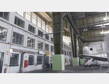 Innenansicht eines historischen Flugzeughangars mit weiß gekachelten Wänden, großen Metallträgern und zahlreichen Fenstern. Im Vordergrund ist das Heck eines silbernen Flugzeugs sichtbar, daneben eine Treppe für den Einstieg; eine WC-Beschilderung ist ebenfalls erkennbar.