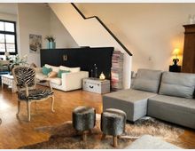 Wohnzimmer mit gemütlicher Einrichtung, einschließlich grauer Ecksofas, einer weißen Couch mit farbigen Kissen, einem Zebra-gestreiften Sessel und einem hellen Holzboden. Ein Treppenaufgang mit schwarzer Akzentwand, ein hoher Bücherstapel und eine Stehlampe ergänzen das moderne Design.