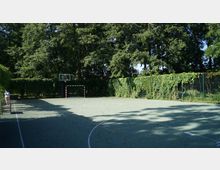 Ein Sportplatz mit einem Basketballkorb und einem Fußballtor, umgeben von hohen Bäumen und dicht bewachsenen Hecken. Der Boden des Platzes besteht aus einem kunststoffbeschichteten Belag mit Spielfeldmarkierungen.