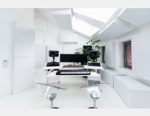 Ein modern eingerichtetes Tonstudio mit weißem Interieur, einem großen Schreibtisch mit einem MIDI-Keyboard, zwei Studiomonitoren und einem Ultrawide-Monitor. Der Raum verfügt über schallabsorbierende Wand- und Deckenpaneele, Oberlichter und Fenster, die einen Blick auf eine Backsteinfassade bieten.