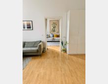 Wohnzimmer mit hellem Holzboden und minimalistischer Einrichtung. Im Vordergrund ein graues Sofa und ein weißer Schrank; durch eine Türöffnung ist ein weiterer Raum mit gepolsterter Sitzbank, bunten Kissen und großem Pflanzenbild an der Wand zu sehen.