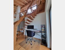 Moderner Arbeitsbereich unter einer offenen Holztreppe mit freitragenden Stufen und Metallgeländer. Der Raum hat eine Holzdecke, einen Schreibtisch mit Monitor und Bürostuhl sowie ein Regal an der Wand.