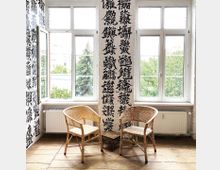 Zwei Rattanstühle stehen in einem hellen Raum mit großen Fenstern, durch die Tageslicht einfällt. Die Wände sind mit großflächiger Kalligrafie in asiatischen Schriftzeichen dekoriert, und der Boden besteht aus Holzplanken.