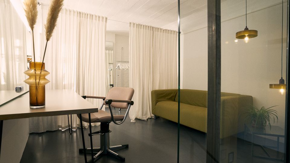 Helles, modernes Innenraumstudio mit halbtransparenter Glaswand, weißem Vorhang als Raumteiler und dunkelgrauem Boden. Links stehen ein Tisch mit Vase und ein verstellbarer Stuhl, rechts ein grünes Sofa mit hängenden Lampen und einer kleinen Pflanze.