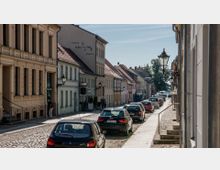 Eine gepflasterte Straße in einer historischen Altstadt mit Reihenhäusern im klassischen Stil, teils mit Ladenfronten und Gasthäusern. Autos parken am Straßenrand, und vereinzelt sind Fußgänger unterwegs; Laternen im traditionellen Design säumen die Straße.