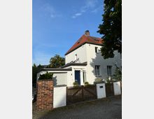 Einfamilienhaus mit weißer Fassade und rotem Ziegeldach, vorne mit blauem Eingang und angebauter Garage. Davor stehen ein niedriger Holzzaun sowie gemauerte Pfeiler, umgeben von Bäumen unter blauem Himmel.