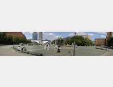 Weitläufiger Platz in einer Stadt mit dem Neptunbrunnen im Zentrum, umgeben von Bäumen, Sitzbänken und historischen sowie modernen Gebäuden; im Hintergrund ist der Fernsehturm und das Rote Rathaus sichtbar. Menschen spazieren und verweilen auf dem gepflasterten Platz.
