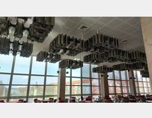 Modernes Café oder Veranstaltungsraum mit großen Fensterfronten, die viel Tageslicht hereinlassen. Auffällige Deckeninstallationen aus röhrenförmigen Konstruktionen und rote Polsterstühle prägen den Innenraum.