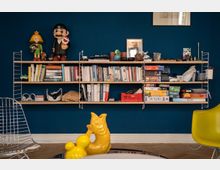 Wall-mounted shelving on a dark blue living-room wall filled with books, board games, and small decorative objects, with framed artwork above. Modern seating includes a wire chair and a yellow chair, and a low table holds a yellow sculptural piece in the foreground.